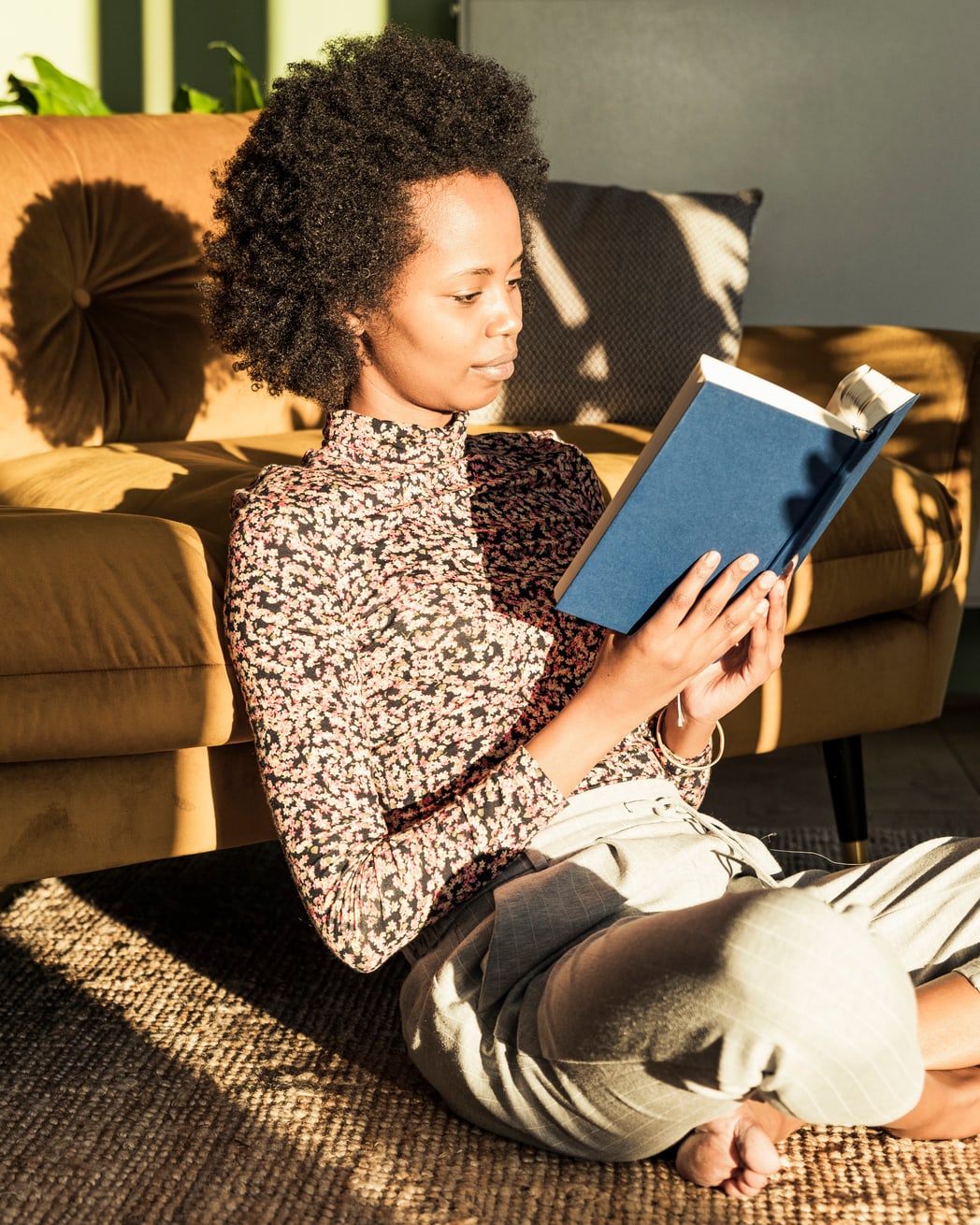 Una mujer está sentada en el suelo, apoyada en un sofá de cuero. Está leyendo un libro en una habitación soleada.